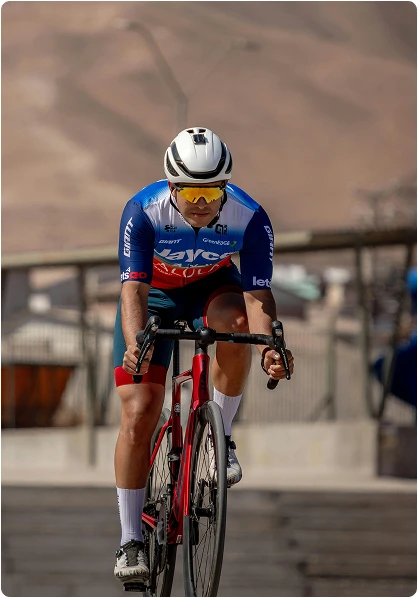 A cyclist wearing a white helmet and blue outfit rides a red bike on a sunny day, focused and intense. Desert hills form a blurred background.
