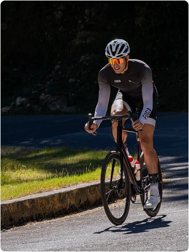 A cyclist in a black and white outfit rides on a sunny road, focused and determined. Greenery is visible at the roadside, casting shadows on the path.