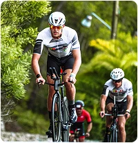Cyclists wearing helmets and matching outfits ride on a road surrounded by lush vegetation. The leading cyclist shows determination and focus.