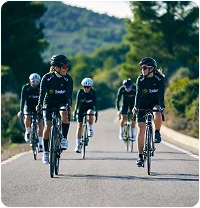 A group of cyclists wearing black outfits rides on a scenic road surrounded by lush vegetation. The atmosphere is energetic and focused under a clear sky.
