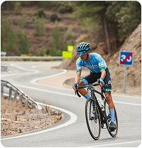A cyclist in a blue jersey and helmet descends a winding mountain road, focused. In the background, trees and road signs can be seen.