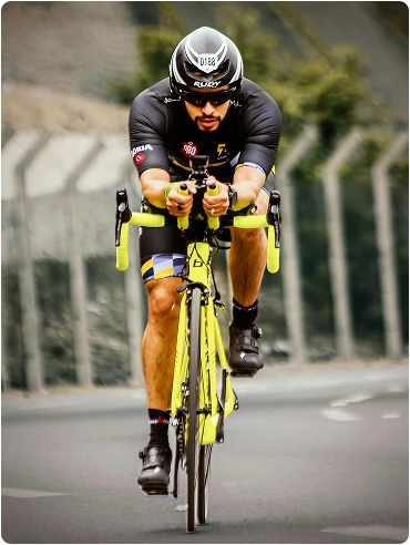 A cyclist dressed in dark attire and a helmet rides on a road with a bright yellow bike. The background is blurred, featuring a fence and greenery, creating an impression of speed and focus.