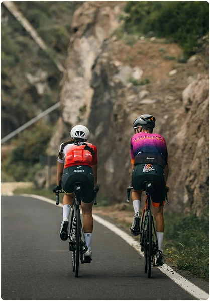 Two cyclists climb a winding road flanked by rocky cliffs and greenery. They wear colorful jerseys and helmets, exuding concentration and determination.